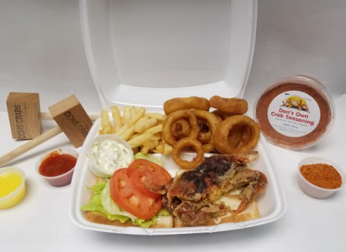 A takeout box with fried chicken, fries, onion rings, tomatoes, lettuce, and sauces, alongside crab seasoning.