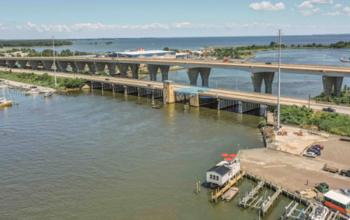 Aerial view of a bridge over water, with boats docked and a marina in the foreground under a clear blue sky.