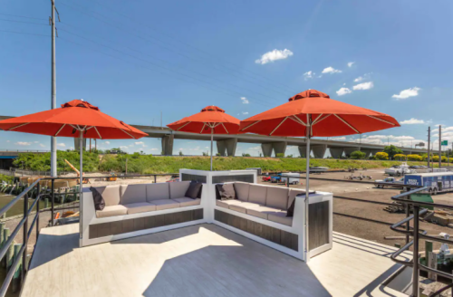 A rooftop lounge area with red umbrellas and seating, overlooking a highway and green landscape under a clear blue sky.