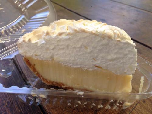 A slice of lemon meringue pie with a golden crust and fluffy white meringue topping in a clear plastic container.