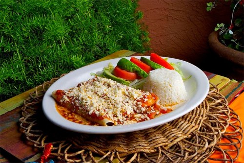 A plate of enchiladas topped with cheese, served with rice and fresh vegetables on a woven mat.