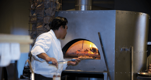 A chef in a white coat uses a long tool to place food into a large, wood-fired oven. Flames are visible inside.