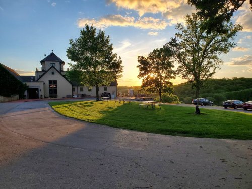 A scenic view of a driveway leading to a building, with trees and a sunset in the background. Tables are set up on the grass.