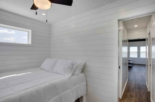 Cozy bedroom with a white shiplap wall, a bed with white bedding, and a ceiling fan, leading to another room.