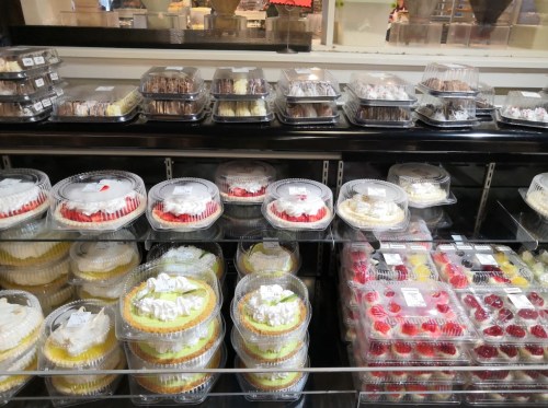 A display case filled with various cakes and desserts in clear plastic containers.