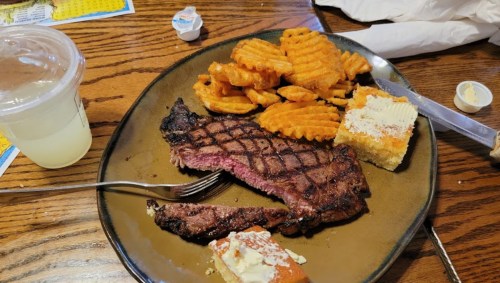 A plate with a grilled steak, waffle fries, cornbread, and a drink on a wooden table.