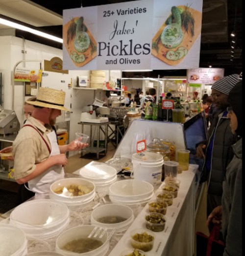 A vendor at a market offers samples of pickles and olives, with various containers displayed on the counter.