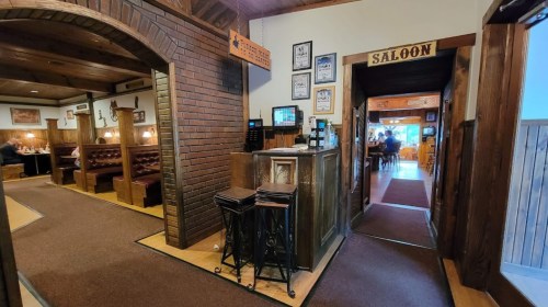 Interior of a rustic restaurant with wooden decor, a bar area, and a sign for "SALoon" leading to a dining area.