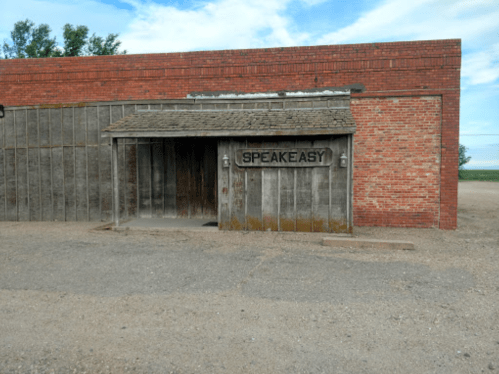 A rustic building with a wooden entrance and a sign reading "SPEAKEASY," set against a blue sky.