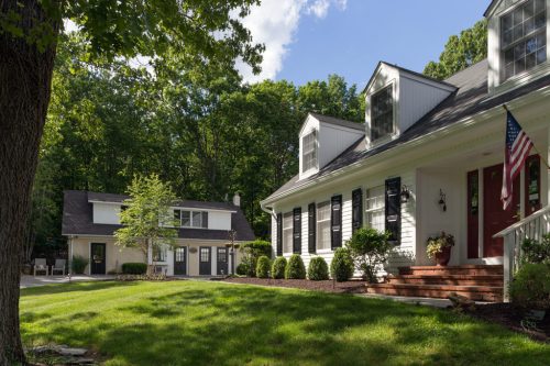 A charming white house with black shutters and an American flag, surrounded by greenery and a separate garage.