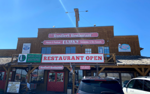 Exterior of Rustler's Restaurant with signs for groceries, dry goods, and an open restaurant. Clear blue sky above.