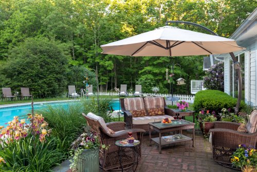 A cozy patio with seating, an umbrella, and a pool surrounded by lush greenery and colorful flowers.