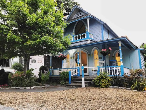 A charming blue house with a porch, surrounded by greenery and flowers, featuring decorative architectural details.