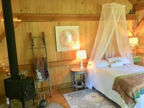 Cozy bedroom with wooden walls, a bed draped in a canopy, a vintage lamp, and a wood stove. Warm and inviting atmosphere.