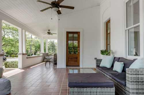 A cozy porch with a wicker seating area, ceiling fans, and a wooden door, surrounded by greenery.
