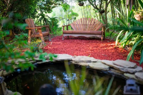 A serene garden scene featuring a wooden bench and chair beside a small pond, surrounded by lush greenery.