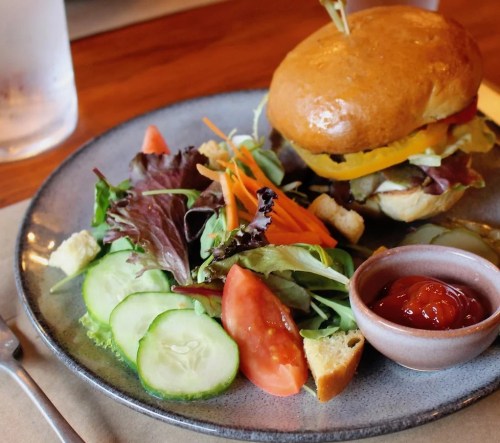 A plate with a burger topped with vegetables, a side salad with cucumbers and tomatoes, and a small bowl of ketchup.