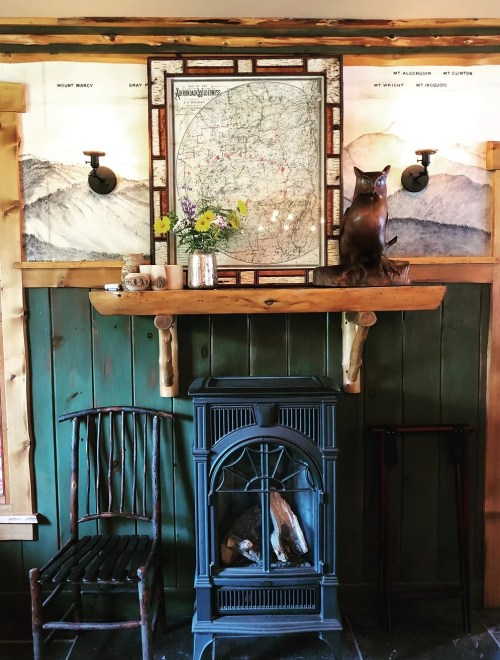 Cozy interior featuring a wooden mantel, vintage map, flowers, an owl statue, and a black stove against green walls.