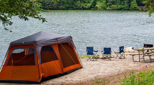 A tent by a calm lake, with camping chairs and a picnic table nearby, surrounded by trees.