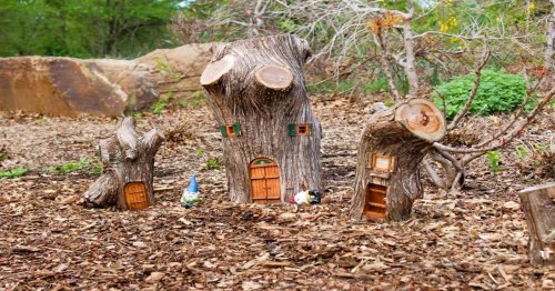 Whimsical tree stump houses with small doors, surrounded by wood chips and greenery. A gnome stands nearby.