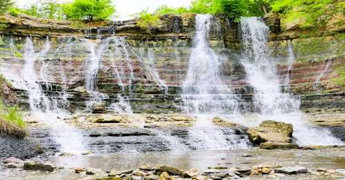 A scenic waterfall cascading over layered rock formations, surrounded by lush greenery and a calm river below.