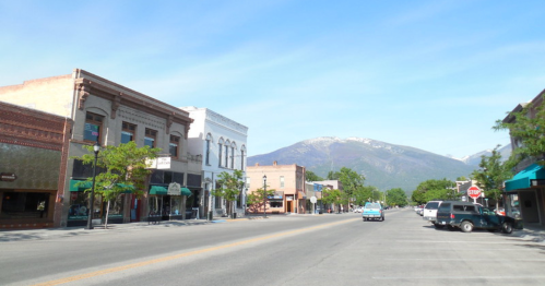 A quiet street lined with historic buildings, trees, and mountains in the background under a clear blue sky.