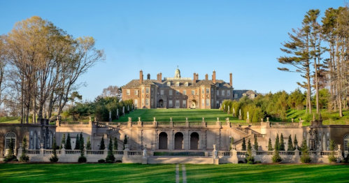A grand mansion with a manicured lawn, surrounded by trees and elegant stone architecture under a clear blue sky.