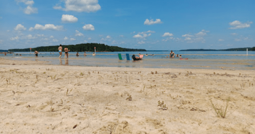 A sandy beach with people swimming and relaxing in the water under a blue sky with fluffy clouds.