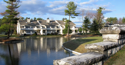 A serene view of a pond with stone walls and modern buildings surrounded by trees under a blue sky.