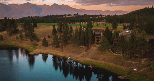 A serene landscape featuring a lake, trees, and mountains at sunset, with cabins along the shore.