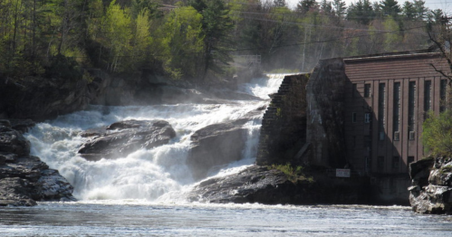 A waterfall cascades over rocks beside an old stone building, surrounded by lush greenery and mist.