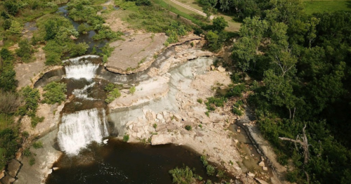 Aerial view of a waterfall cascading over rocky terrain, surrounded by lush greenery and a winding river.