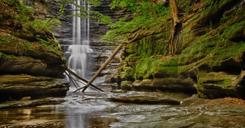 A serene waterfall cascades down rocky cliffs, surrounded by lush greenery and moss-covered stones.