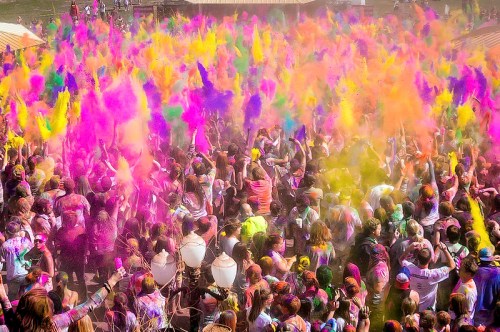A vibrant crowd celebrating with colorful powders in a festival, creating a cloud of pink, yellow, and purple hues.