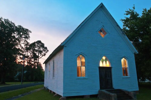 A small white church with arched windows, illuminated at dusk, surrounded by trees and a quiet road.