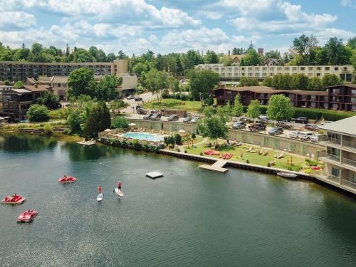 Aerial view of a lake resort with paddleboarders, a pool, and lush greenery under a partly cloudy sky.