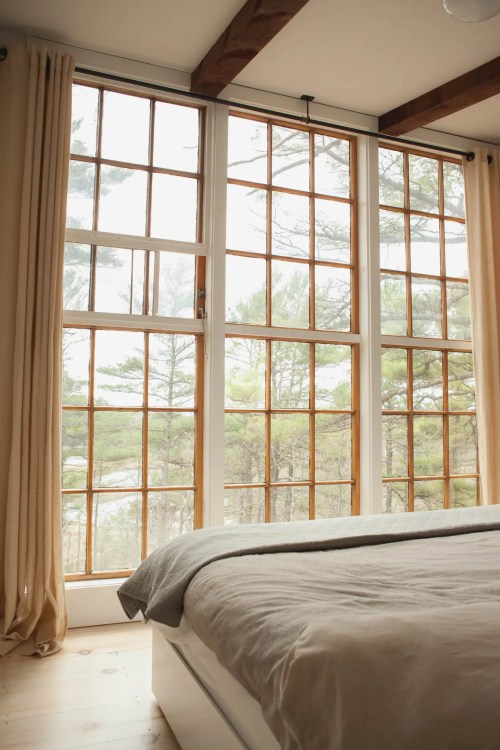 Cozy bedroom with large windows, soft curtains, and a view of trees outside. Natural light fills the space.