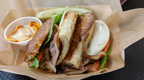 A sandwich with layers of meat, cheese, and vegetables, served with a pickle and a side of dipping sauce.