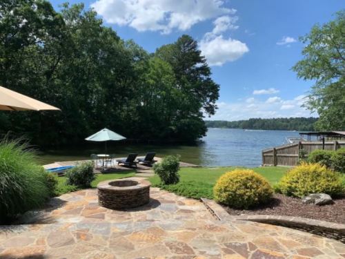 A serene lakeside view with a stone patio, lounge chairs, and a fire pit surrounded by greenery and trees.