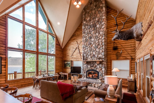 Cozy log cabin interior with large windows, stone fireplace, and rustic decor, overlooking a scenic view.