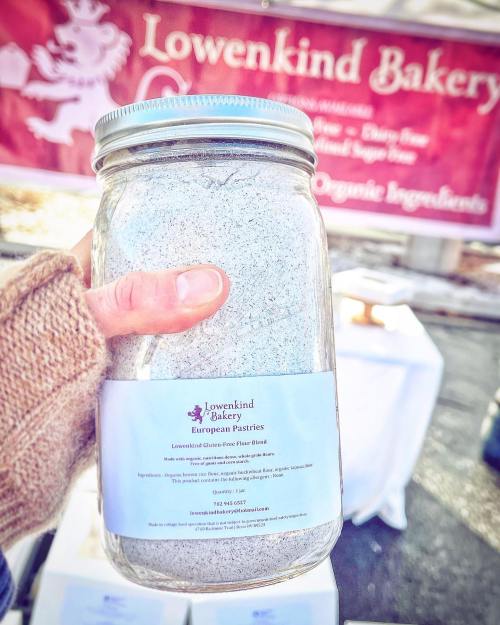 A person holds a jar of Lowenkind Bakery gluten-free flour blend in front of a bakery banner.