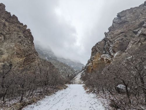 A snowy path winds through a rocky canyon, with clouds hanging low over the mountains in the background.