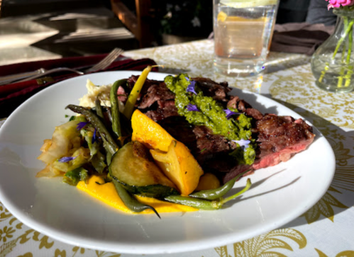 A plate of grilled steak topped with green sauce, served with sautéed vegetables and garnished with edible flowers.