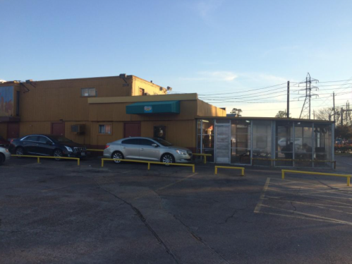 A parking lot with two cars in front of a yellow building and a bus stop shelter under a clear blue sky.