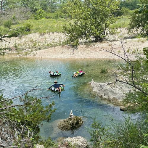 Four people float on inner tubes in a calm river surrounded by greenery and rocky banks on a sunny day.