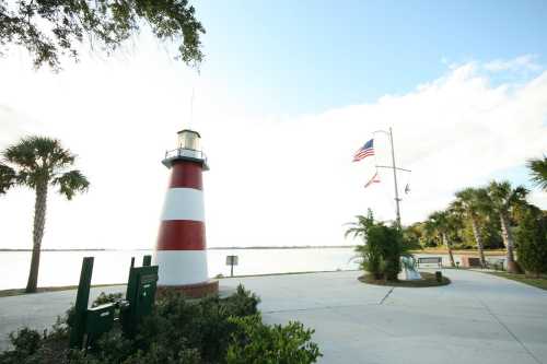 A red and white striped lighthouse near a lake, with palm trees and flags in a scenic park setting.