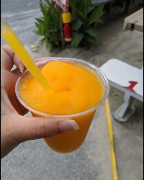 A hand holding a cup of orange slush with a straw, set against a blurred outdoor background.