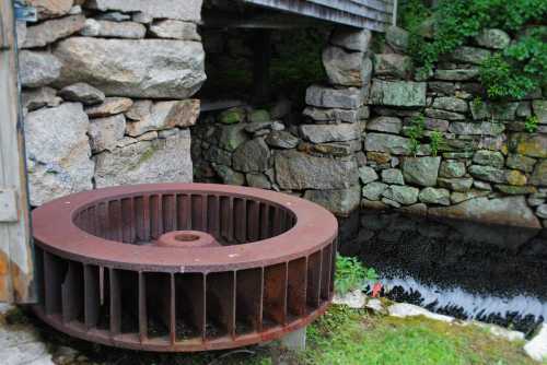 A large, rusted water wheel sits beside a stone wall near a calm body of water, surrounded by greenery.