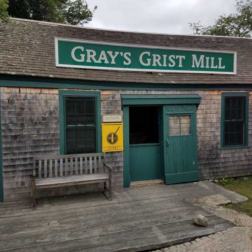 A rustic wooden building with a green door and sign reading "Gray's Grist Mill," surrounded by trees.