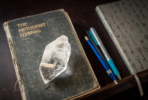 A crystal sits on top of an old hymnal, with pens and a notebook nearby on a wooden surface.
